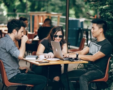 Three coworkers sit at a coffee shop table. The man on the right is typing into an Apple laptop, while the woman in the center leans over to watch the screen. A third man sits on the left, sipping coffee and listening to the other two.